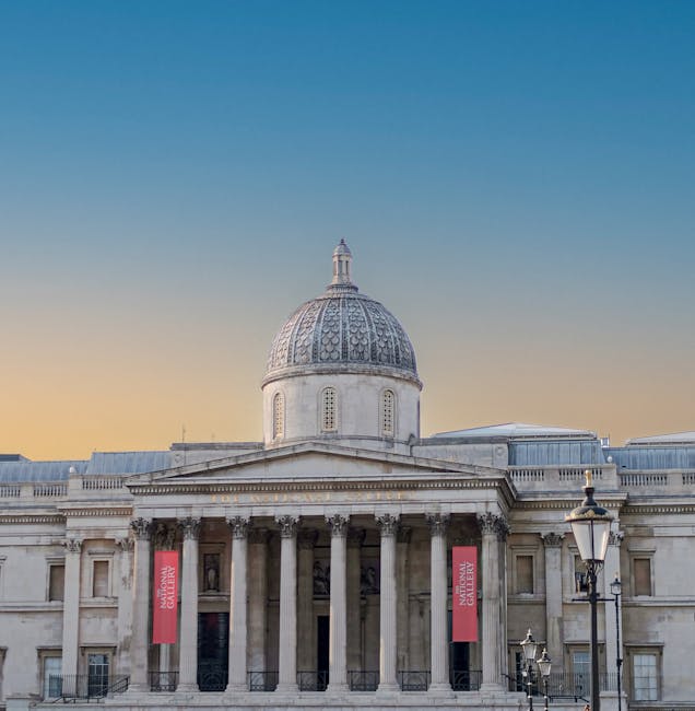 Photograph of a grand neoclassical building with tall, evenly spaced stone columns supporting a triangular pediment decorated with sculptures. The facade is made of light-colored stone, and a flag is
