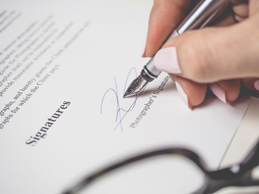 Close-up of a person's hand holding a silver pen and signing a document titled 'Signatures', with a printed paragraph visible in the background. The hand has neatly manicured nails with a light pink p