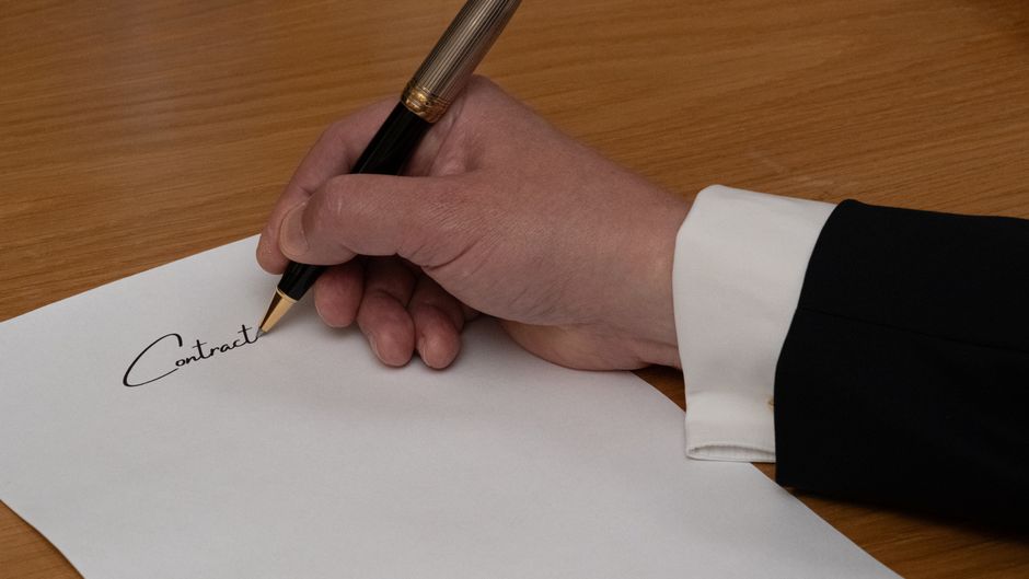 Close-up of a person's hand holding a silver pen and signing a document titled 'Signatures', with a printed paragraph visible in the background. The hand has neatly manicured nails with a light pink p
