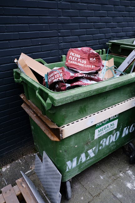 A black, cylindrical public trash bin with a domed lid, situated on a cobbled street in an urban area. The top of the bin is cluttered with paper cups, paper and plastic waste, and empty glass bottles
