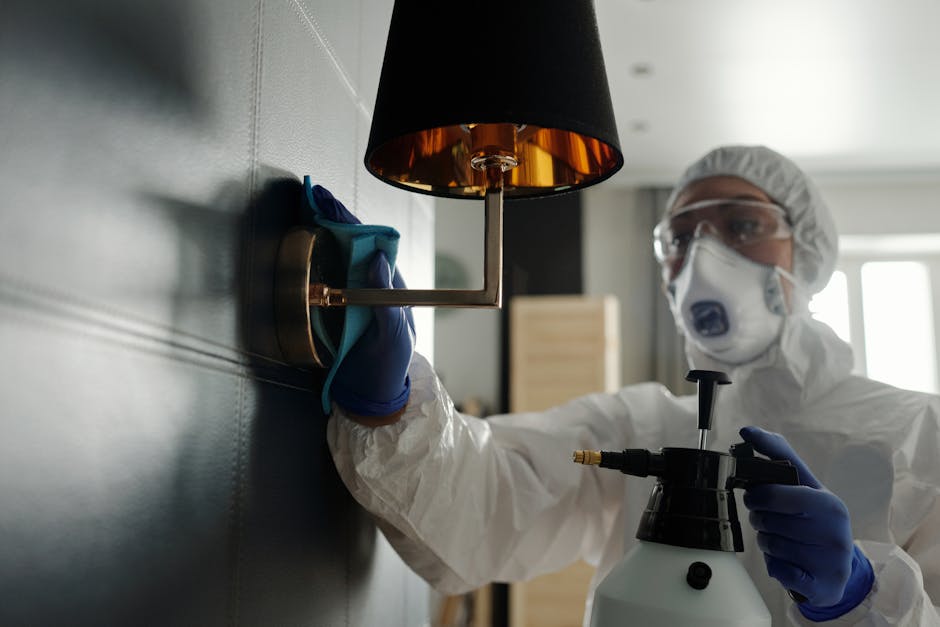 A professional cleaner dressed in white protective suits, wearing gloves and a face mask, kneeling on a wooden floor while disinfecting a surface with a cloth. In the foreground, there is a spray bott