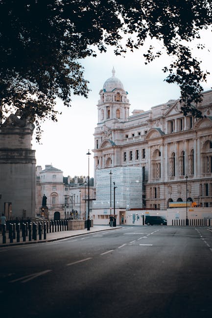 A street scene in Russell Square featuring a historic building with intricate stonework, domed towers, and ornate windows, partially shaded by the leafy branches of large trees overhead. The road is e
